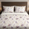 Front close view of ivory bedding with scattered purple and lavender pansies, small yellow flowers and delicate gray-green leafy stems on the cover and matching pillows.