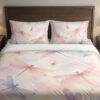 Front close view of bedding printed with translucent pink, lavender and blue dragonflies over a soft peach, cream and pink watercolor ground on the cover and matching pillows.