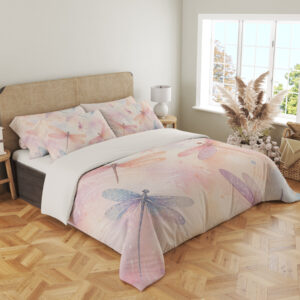 Angled full-bed view of bedding printed with translucent pink, lavender and blue dragonflies over a soft peach, cream and pink watercolor ground, showing the pattern across the cover and pillows.