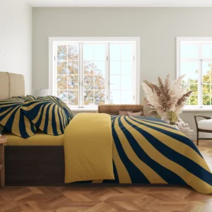 Side view of navy bedding crossed by broad gold-colored ribbons, with printed pillows and a gold-colored fold in a bright windowed room.