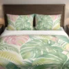 Front view of oversized sage and blush split leaves with pale yellow fronds on a cream duvet cover and matching pillowcases against a dark wooden headboard.