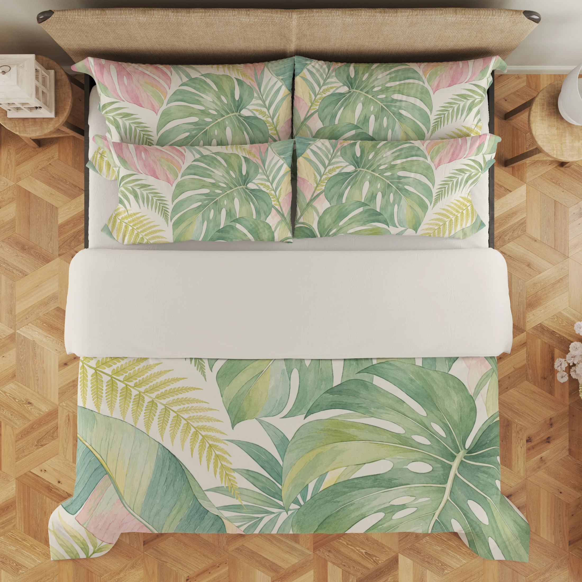 Overhead view of sage and blush split leaves, pale yellow fronds and tropical foliage on the cream duvet cover and styled matching pillows, with a pale fold.