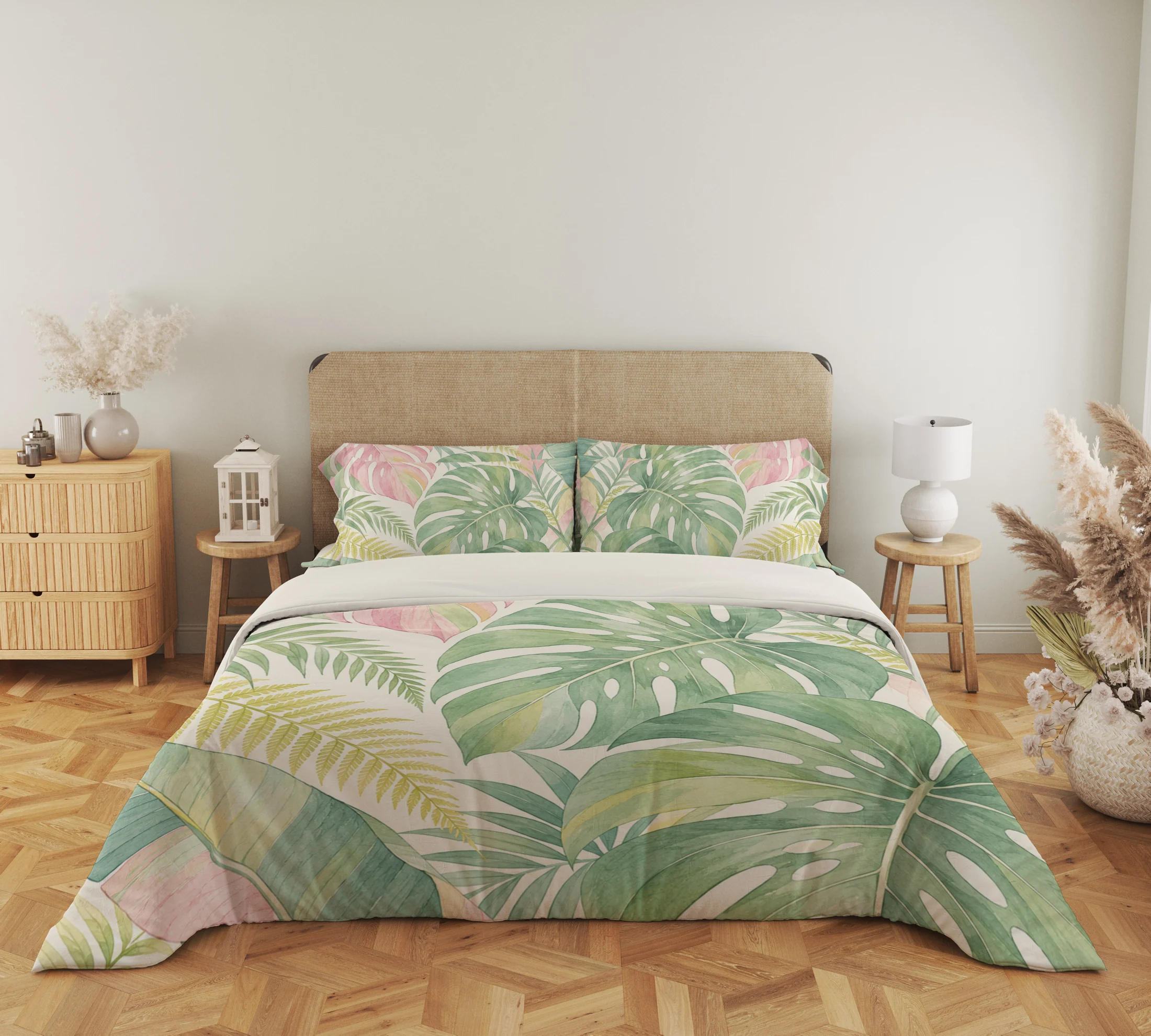 Wide front view of oversized sage and blush tropical leaves with pale yellow fronds on a cream duvet cover and matching pillows, beside a lantern and lamp.