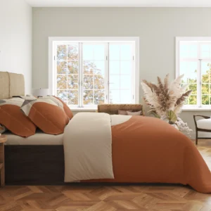 Side view of terracotta and cream abstract bedding with printed pillows and a wide cream fold in a bright room with large windows.